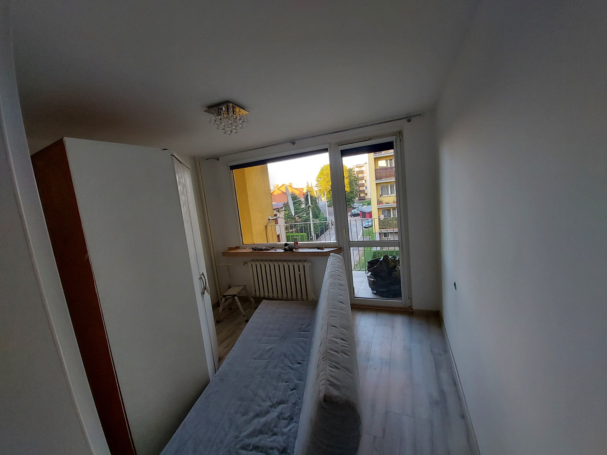 Pusty pokój z białymi ścianami i parkietem, widok na balkon i osiedle za oknem, widoczna szafa z brązowymi wstawkami, mały taboret i grzejnik pod oknem, sufit z lampą kryształową.