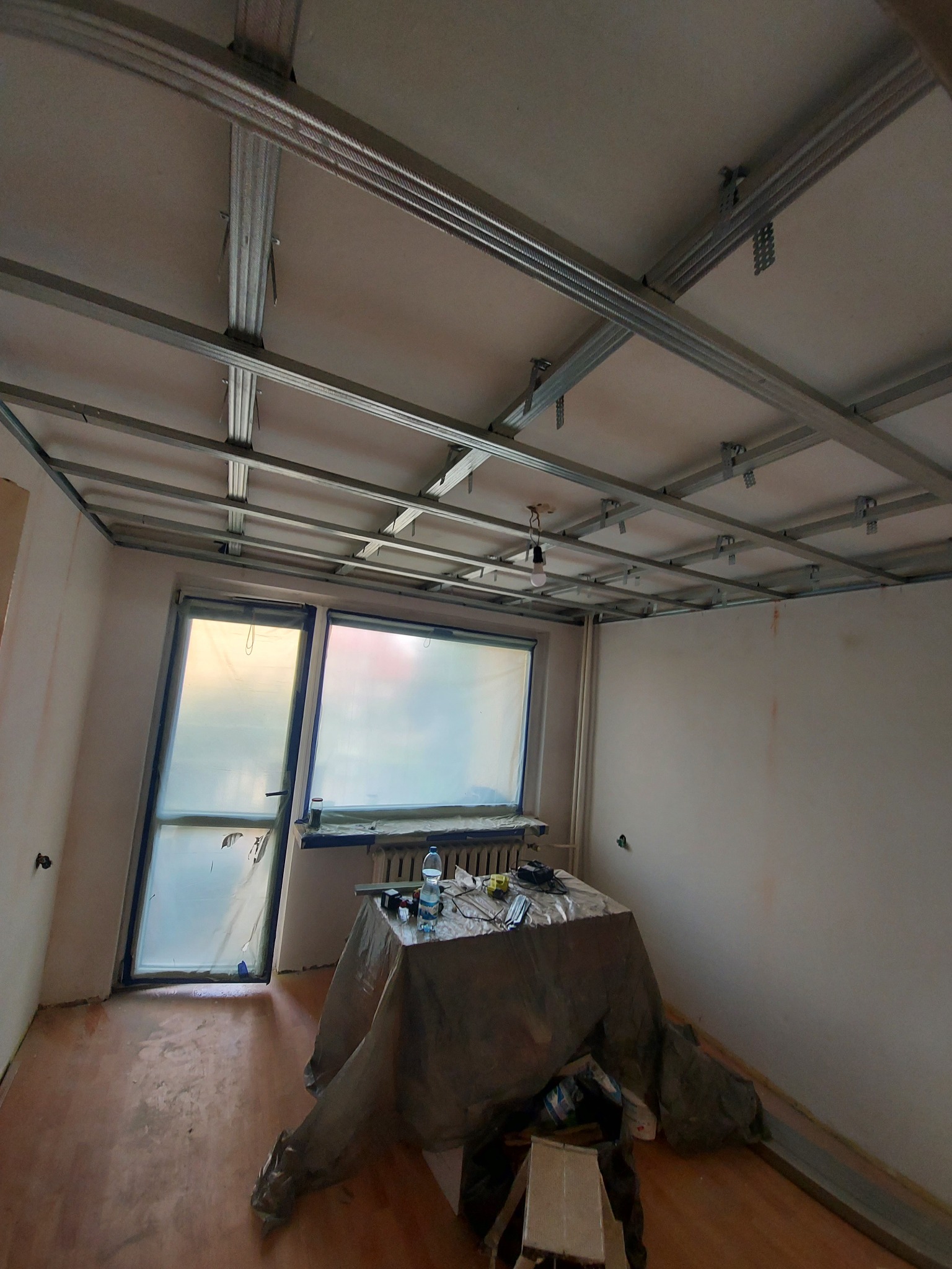 Montaż metalowej konstrukcji sufitu podwieszanego w pokoju, okna zabezpieczone folią, widoczne narzędzia i materiały budowlane.