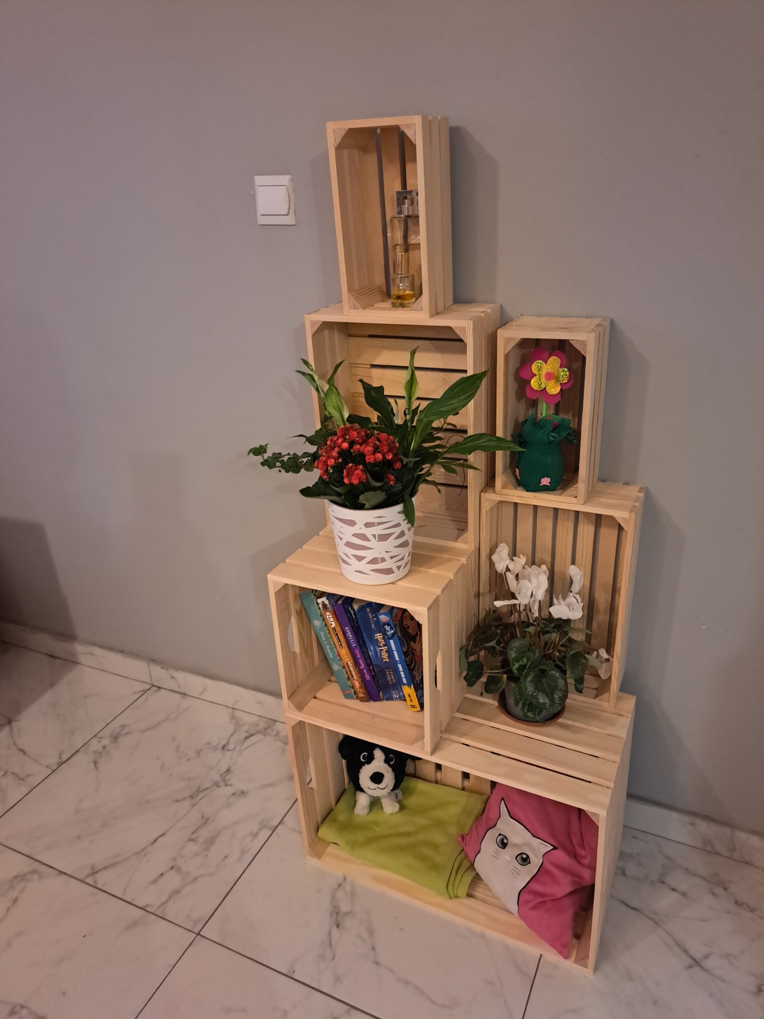 Półka z surowych drewnianych skrzynek ułożonych w piramidę, z dekoracjami: kwiaty doniczkowe, książki, pluszaki, perfumy, na tle szarej ściany i marmurowej podłogi.