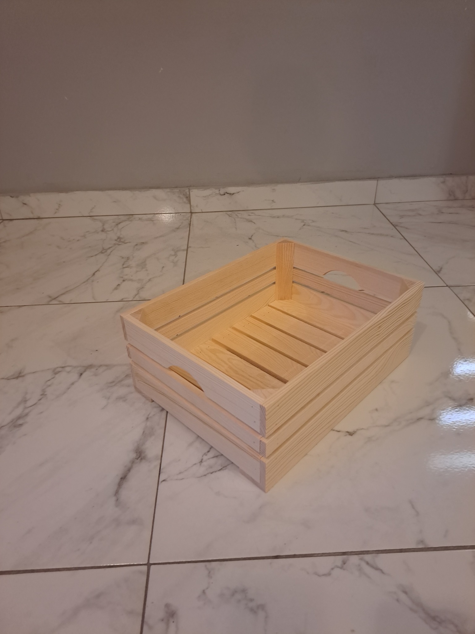 Nielakierowana, jasna, drewniana skrzynka z uchwytami na tle płytek imitujących marmur i szarej ściany.