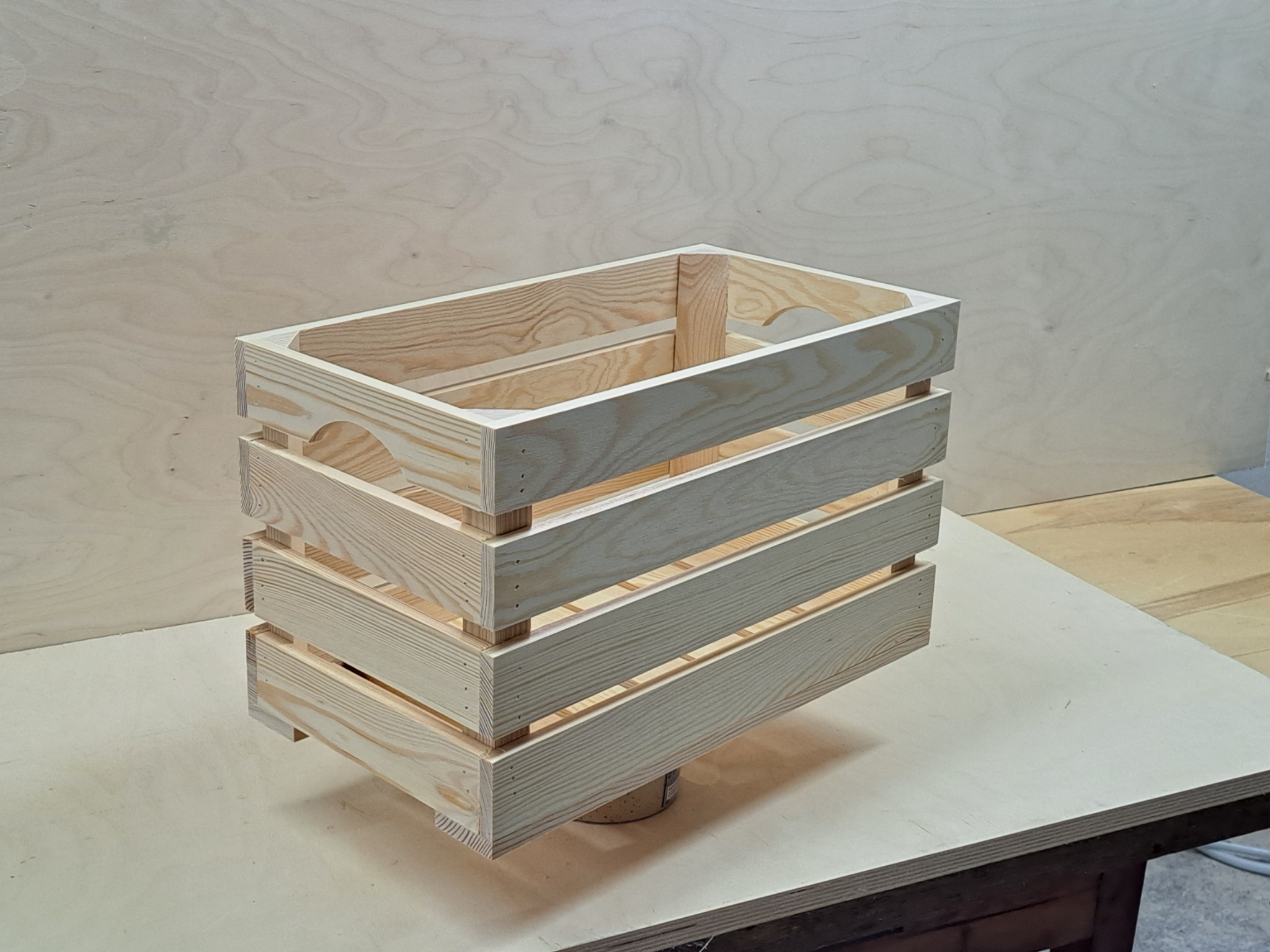 Surowa, drewniana skrzynka z listewek, z zaokrąglonymi uchwytami, na tle sklejki, wsparta na puszce.