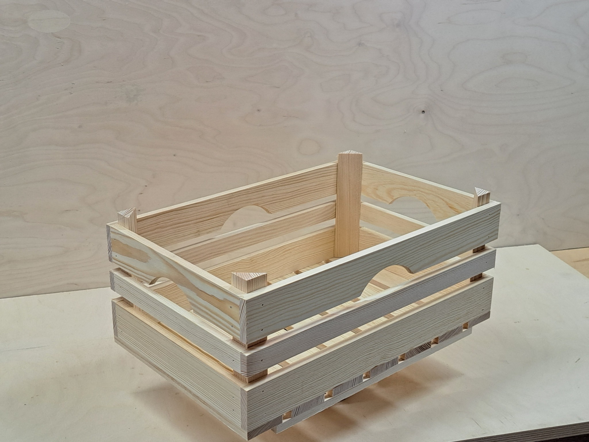 Surowa, drewniana skrzynka z jasnego drewna sosnowego, z uchwytami w kształcie półksiężyca, na tle płyty z forniru drewnianego, ukazująca detale konstrukcyjne i słoje drewna.