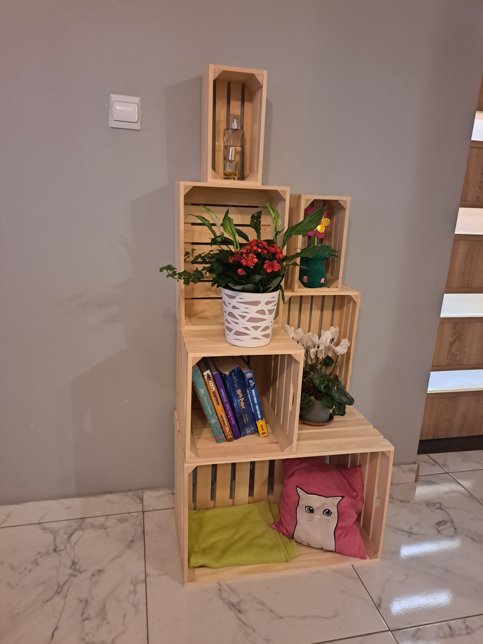 Kaskadowy regał z jasnego drewna, zbudowany z drewnianych skrzynek, dekorowany kwiatami w doniczkach, książkami i poduszkami na tle szarej ściany i marmurowej podłogi.