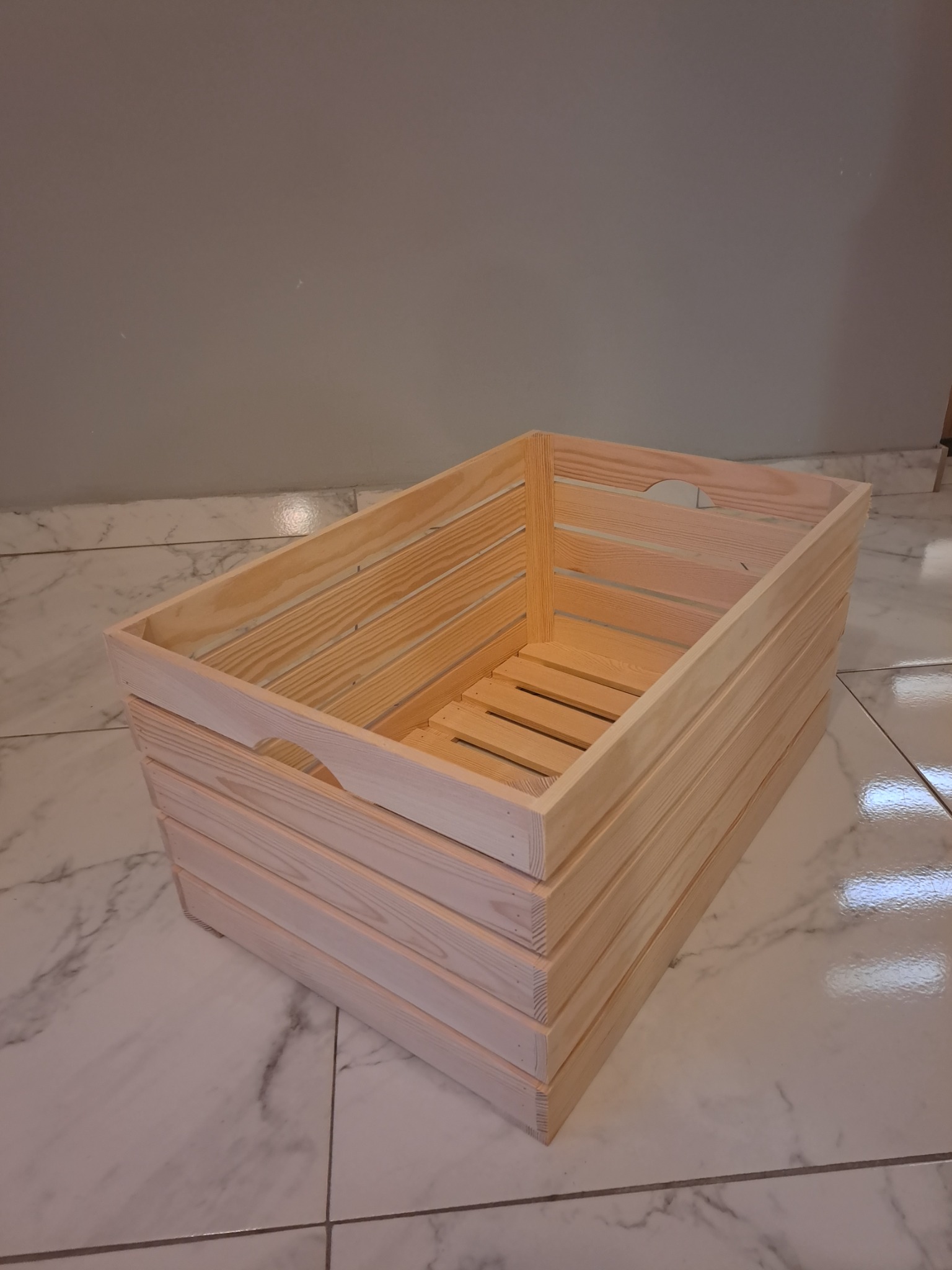 Prosta, surowa skrzynia z jasnego drewna, z wyciętymi uchwytami, ustawiona na marmurowej podłodze w pomieszczeniu o szarych ścianach.