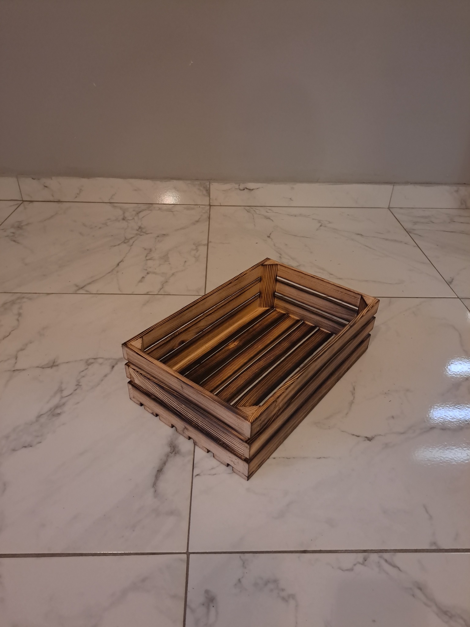 Opalana, drewniana skrzynka z listewek, leżąca na marmurowej podłodze, z widoczną strukturą drewna.