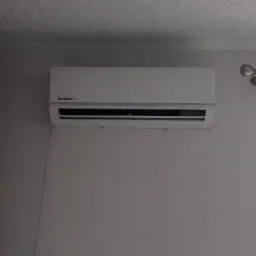 Biały klimatyzator Kaisai Eco zamontowany na szarej ścianie, widoczny kabel zasilający i fragment lampy sufitowej.