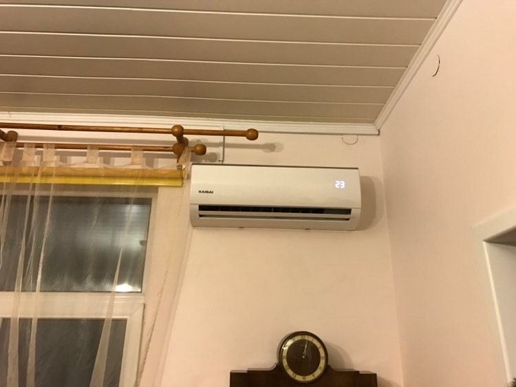 Biały klimatyzator marki Kaisai zamontowany na jasnoróżowej ścianie nad drewnianym zegarem, widoczny fragment okna z firanką i drewniany sufit.