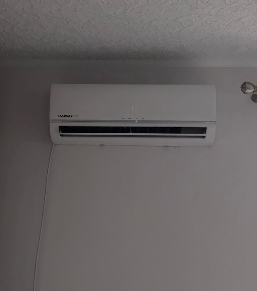 Biały klimatyzator Kaisai Eco zamontowany na szarej ścianie, widoczny kabel zasilający i fragment lampy sufitowej.