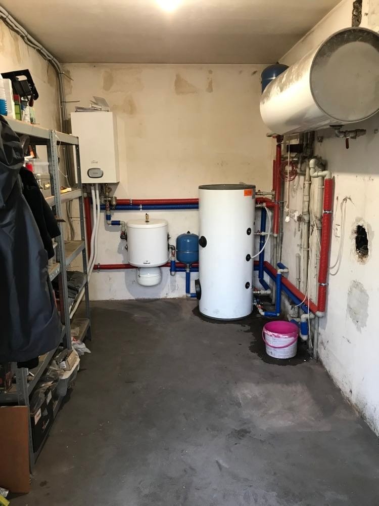Kotłownia z różnymi zbiornikami, piecem gazowym i instalacją hydrauliczną w surowym pomieszczeniu. Widoczna metalowa półka z narzędziami i wiadro z farbą.