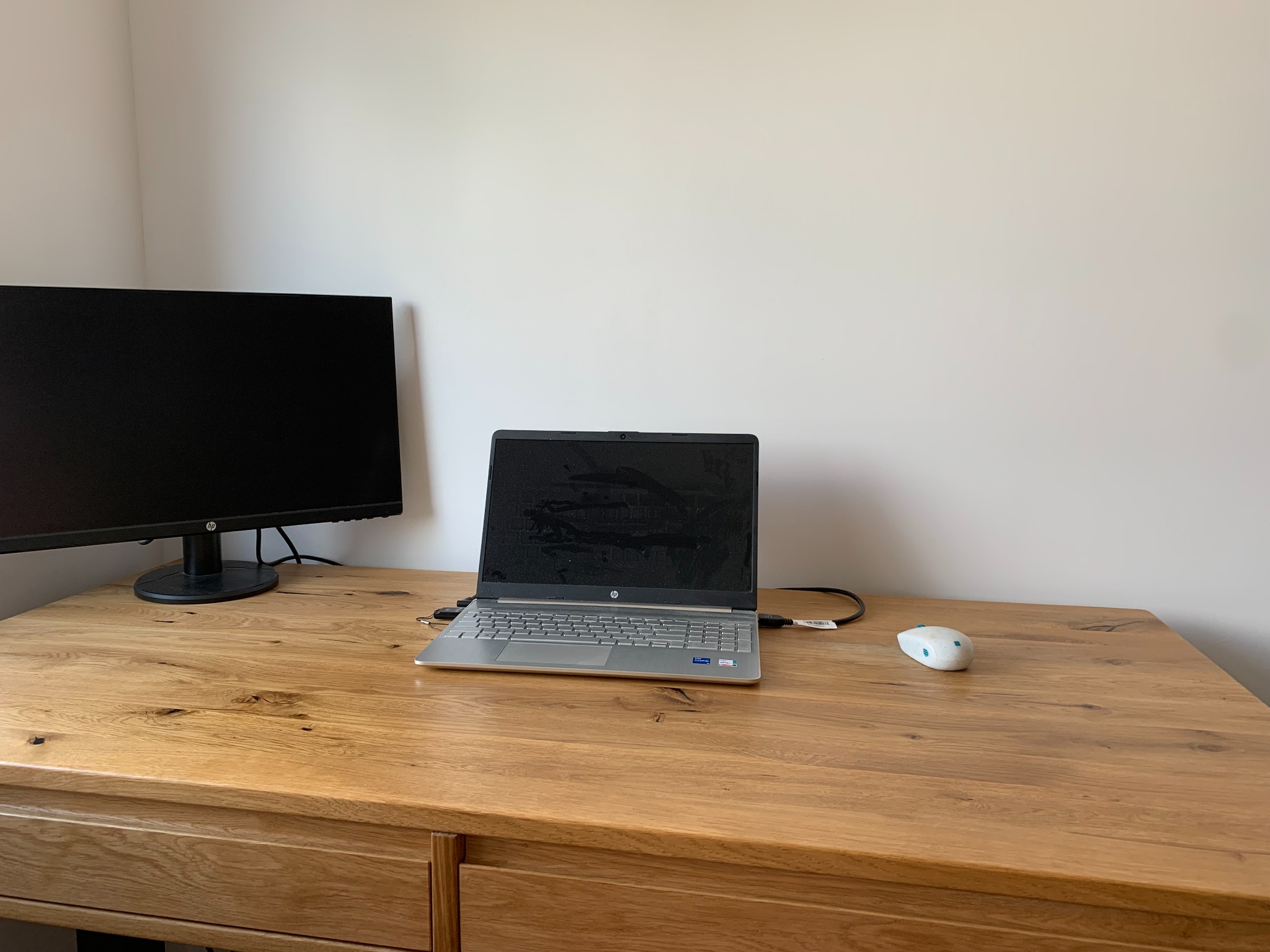 Biurko z laptopem HP, monitorem i myszką na tle jasnej ściany. Drewniany blat biurka z widocznymi słojami. Minimalistyczne, domowe biuro.