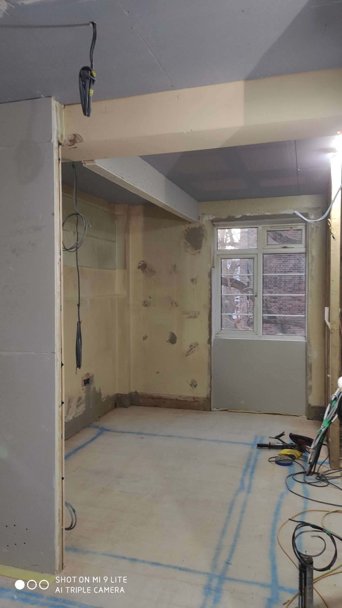 Wnętrze w trakcie remontu: ściany przygotowane do malowania z widocznymi śladami po naprawach, sufit częściowo podwieszany, niebieskie linie wyznaczające obszar na podłodze, narzędzia i kable...