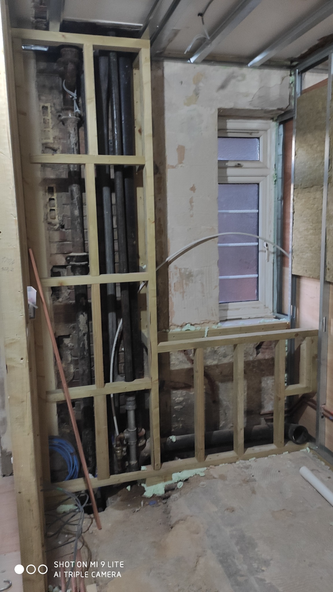 Demontaż starej instalacji wodnej i kanalizacyjnej w trakcie remontu. Widoczne rury w drewnianej ramie, fragment okna i ściany z resztkami tynku.