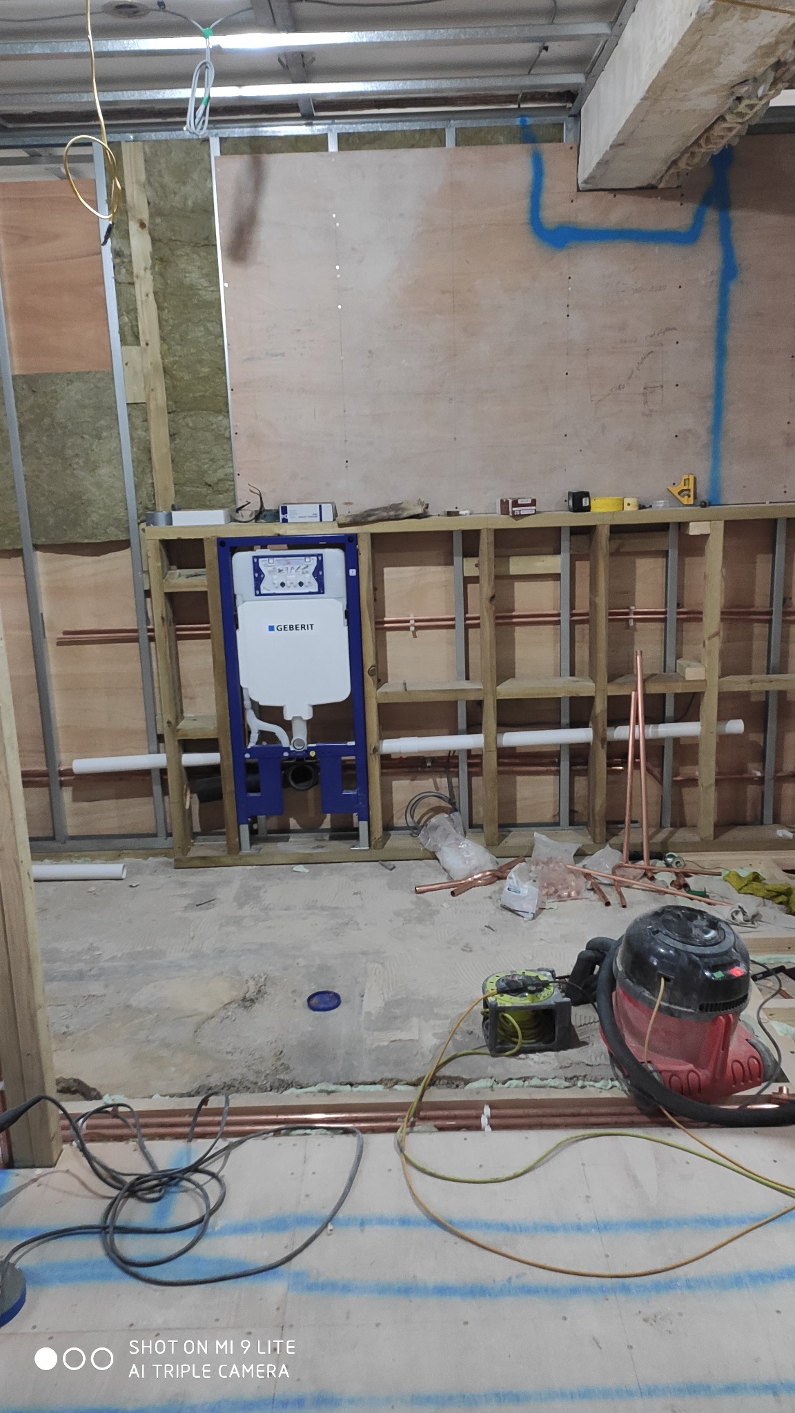 Stelaż podtynkowy Geberit zamontowany w drewnianej konstrukcji na tle surowej ściany z izolacją, widoczne rury miedziane, narzędzia i odkurzacz przemysłowy na podłodze.