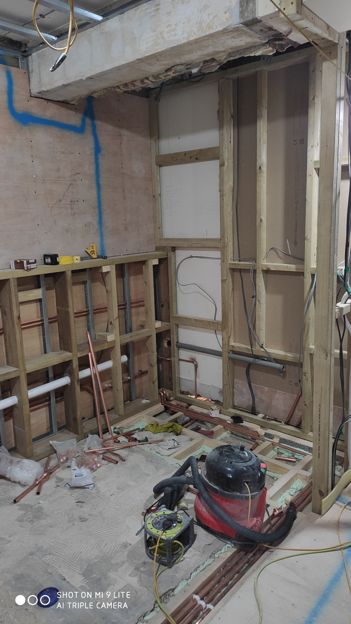 Wnętrze w trakcie remontu z widoczną konstrukcją drewnianą ścian, instalacją hydrauliczną z miedzianych rur, odkurzaczem przemysłowym i przedłużaczem na podłodze.