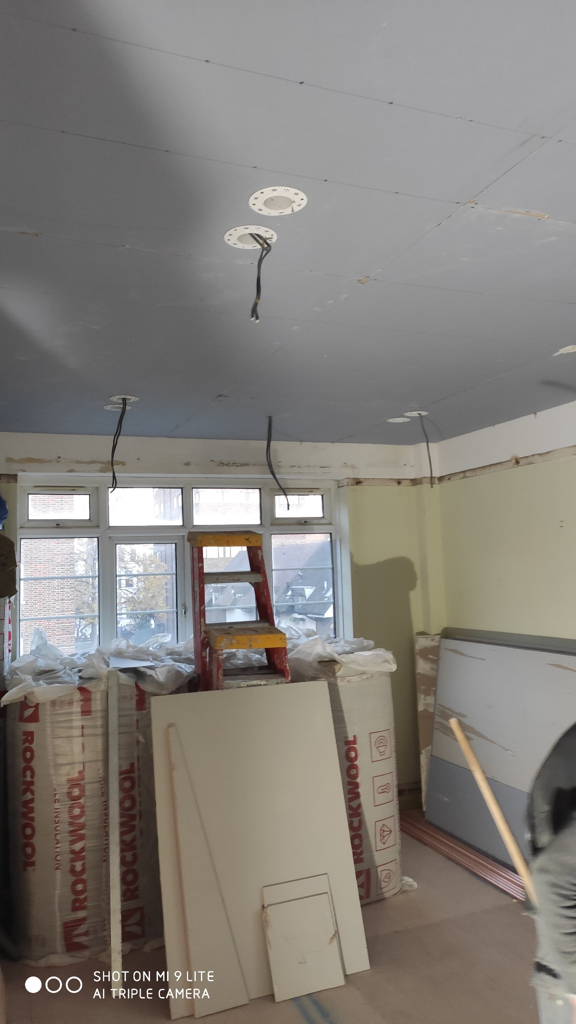 Pokój w trakcie remontu: sufit z płyt kartonowo-gipsowych z otworami na oświetlenie, izolacja Rockwool, drabina malarska, okno z widokiem na budynki.