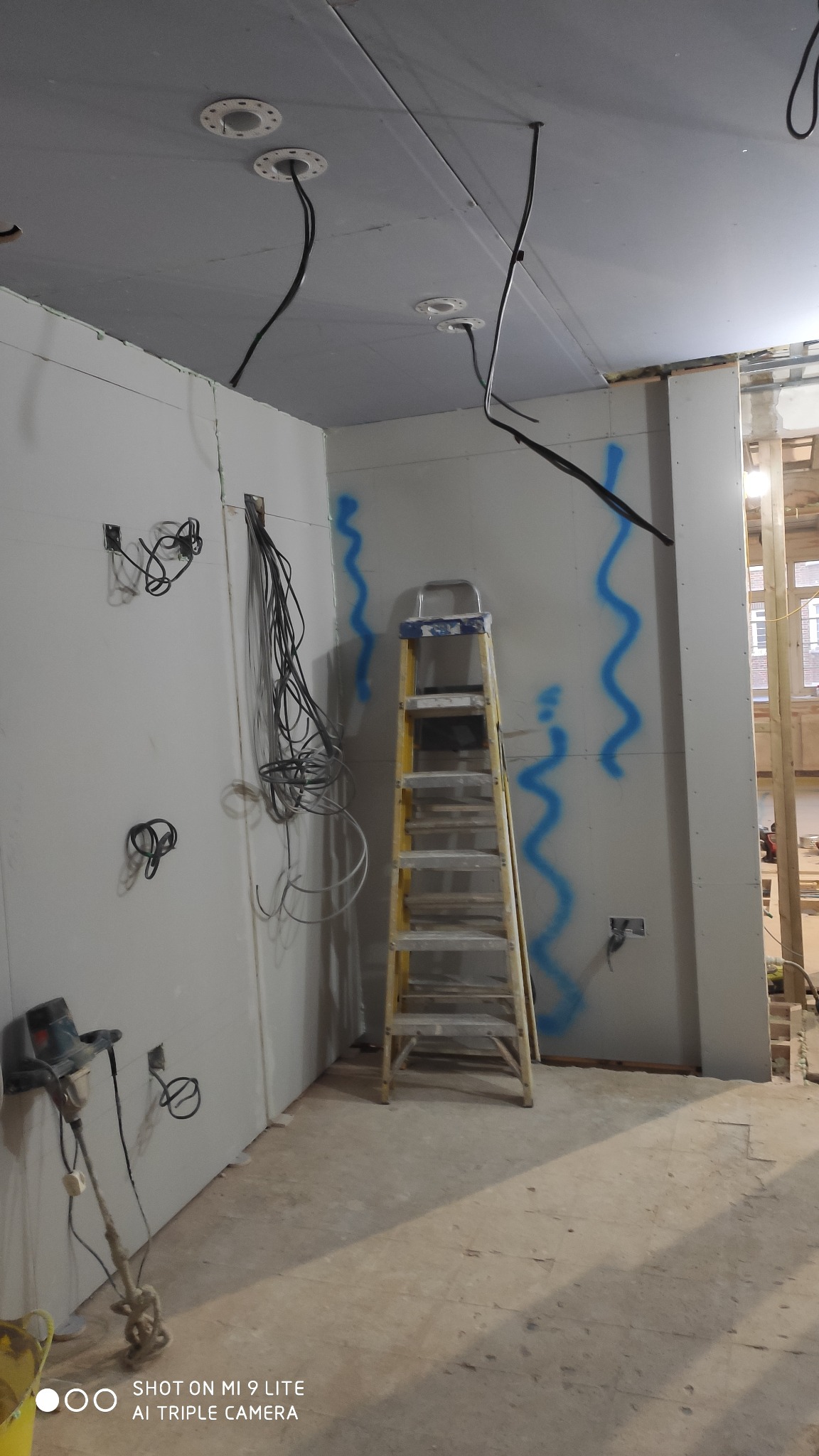 Wnętrze w trakcie remontu z widoczną ścianą z płyt kartonowo-gipsowych, wiszącymi kablami elektrycznymi, żółtą drabiną i śladami niebieskiej farby na ścianie