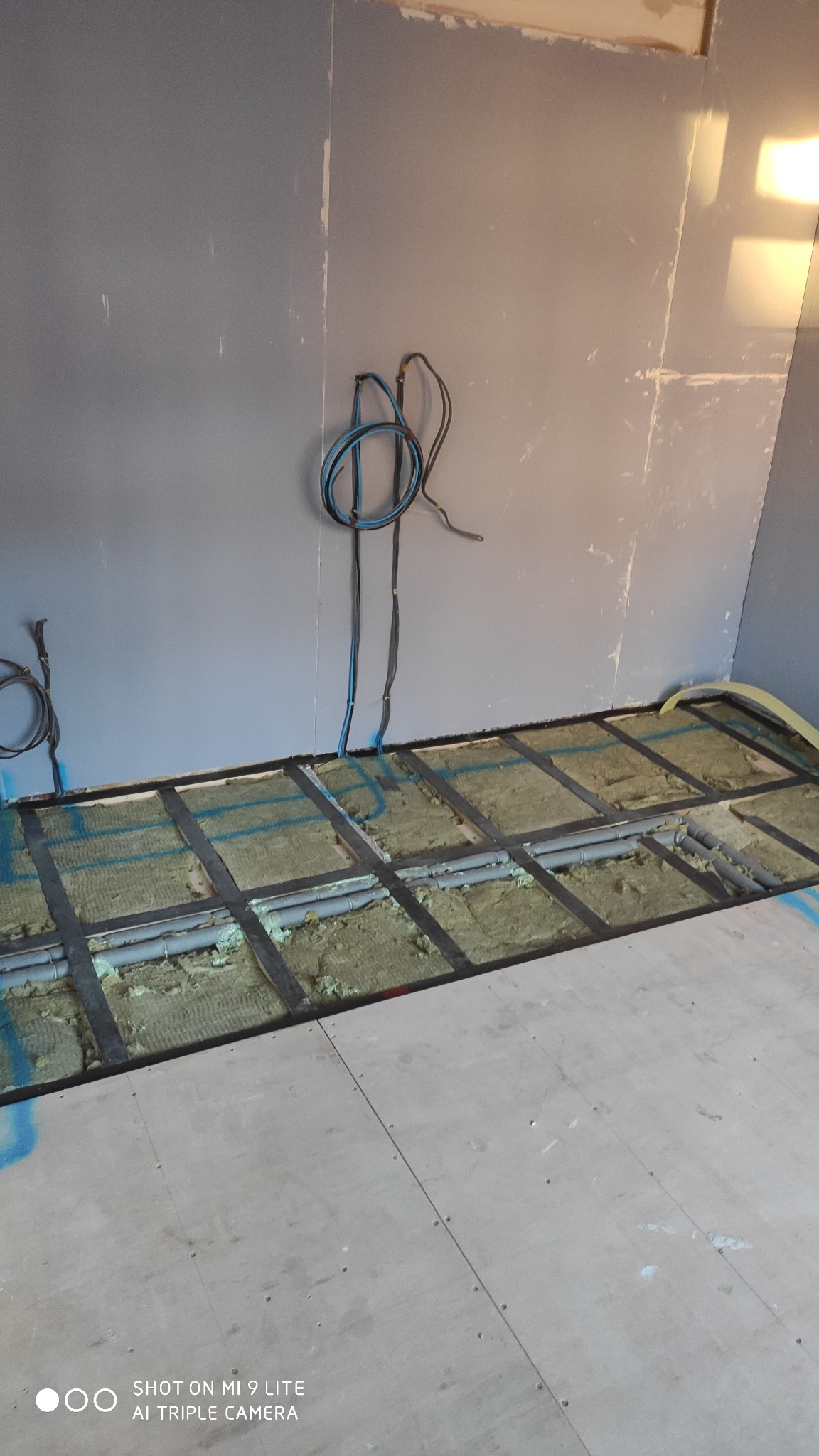 Podłoga z widocznym systemem ogrzewania podłogowego w trakcie montażu, ocieplona wełną mineralną, z częściowo ułożonymi płytami OSB, na ścianie widoczne kable elektryczne.