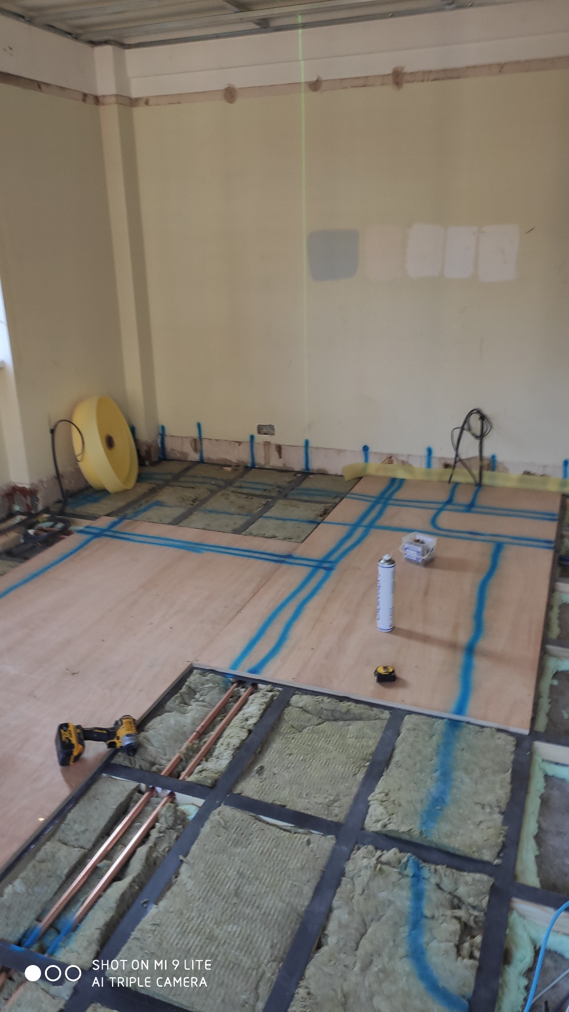 Montaż ogrzewania podłogowego: płyty OSB z wyciętymi kanałami na rury miedziane, wypełnione izolacją, zaznaczone niebieskim markerem, w tle narzędzia i puszka pianki montażowej.