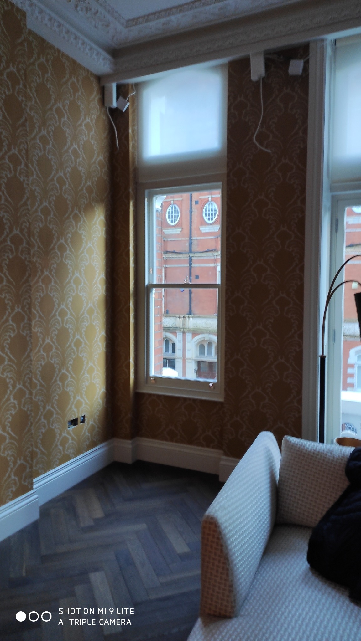 Pokój z wzorzystą tapetą w odcieniach złota, parkietem ułożonym w jodełkę, białymi listwami przypodłogowymi i oknem z widokiem na budynek z czerwonej cegły.