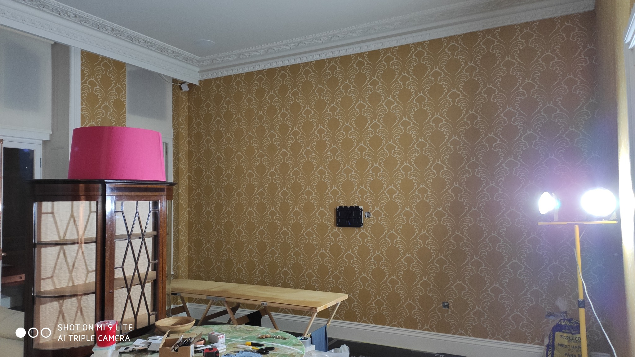 Pokój w trakcie remontu z tapetą w odcieniach złota i bieli, widoczne narzędzia na stole, lampa halogenowa oświetlająca ścianę i ciemny mebel z różowym abażurem.