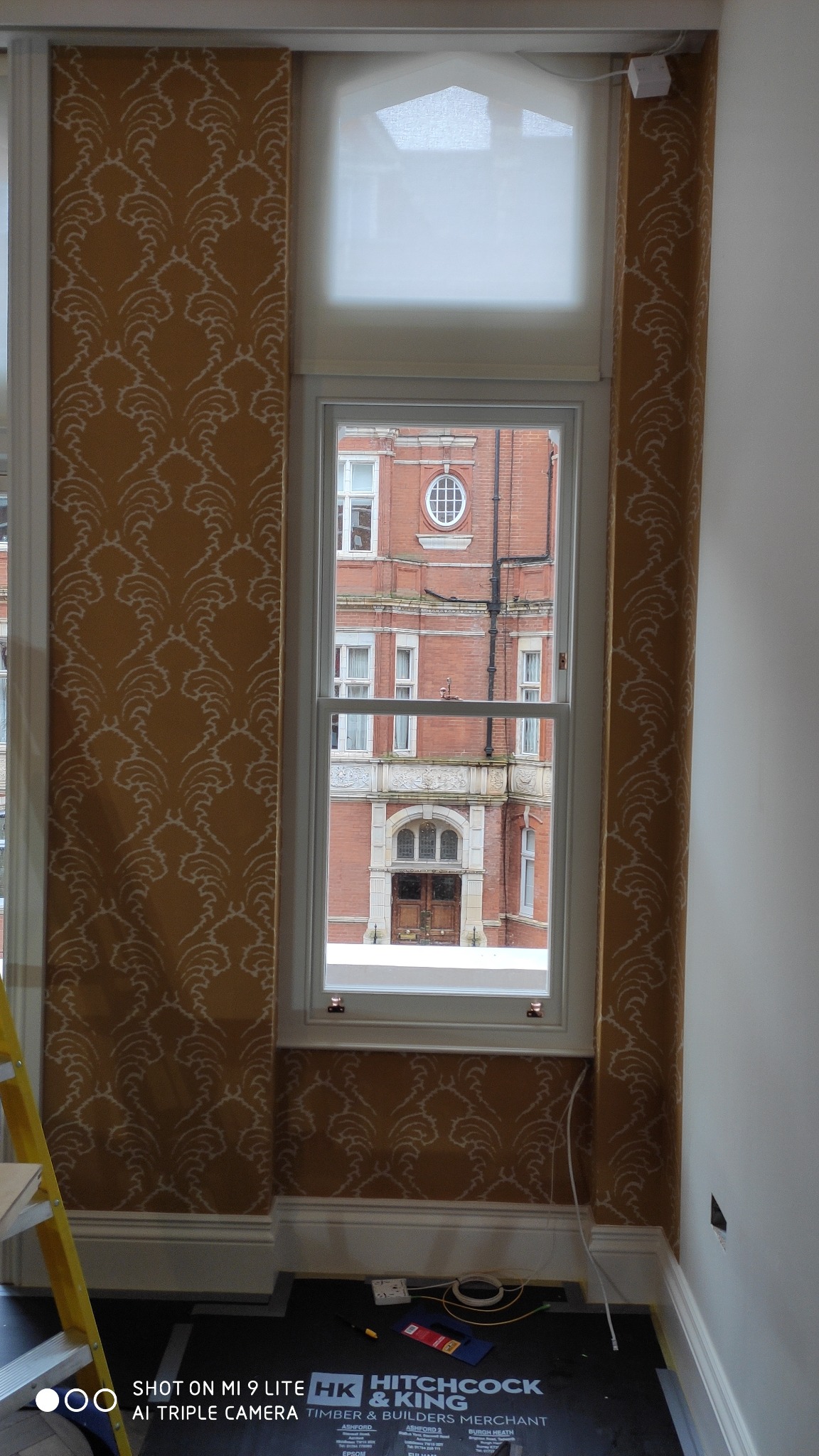 Wnętrze pokoju w trakcie remontu, widoczne tapety w odcieniach złota z wzorem, okno z widokiem na ceglany budynek, fragment drabiny i narzędzia na podłodze zabezpieczonej czarną folią.