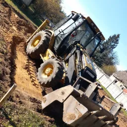 Żółta koparko-ładowarka Caterpillar w trakcie prac ziemnych, widoczny wykop w ziemi, w tle dom i białe ogrodzenie.
