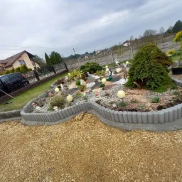 Kształtny rabat z szarej kostki obrzeżowej, wypełniony ozdobnym żwirem, roślinami i dekoracjami ogrodowymi, w tle dom jednorodzinny i samochód.