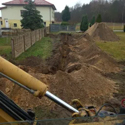 Wykop pod fundamenty lub instalacje widziany z kabiny koparki; w tle dom jednorodzinny i hałdy ziemi.