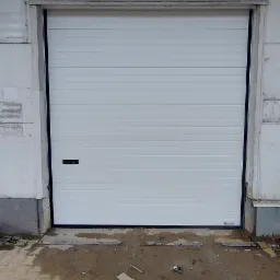 Zamknięta, segmentowa brama garażowa w kolorze białym, z poziomymi przetłoczeniami, zamontowana w surowym, betonowym budynku. Na posadzce widoczne zabrudzenia i drobne śmieci.