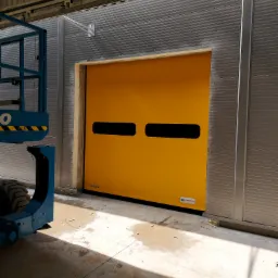 Żółta, szybka brama przemysłowa z dwoma poziomymi okienkami na tle szarej hali, widoczny fragment podnośnika koszowego.