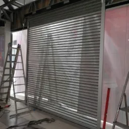 Montaż perforowanej, metalowej rolety w surowym wnętrzu, widoczne drabiny, poziomica i elementy konstrukcyjne budynku.