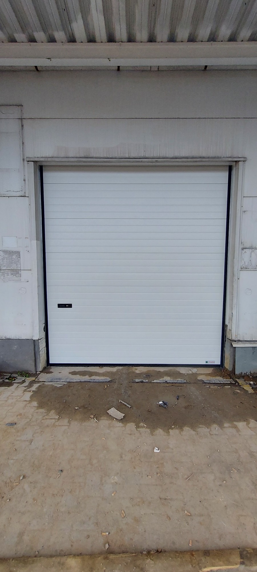 Zamknięta, segmentowa brama garażowa w kolorze białym, z poziomymi przetłoczeniami, zamontowana w surowym, betonowym budynku. Na posadzce widoczne zabrudzenia i drobne śmieci.
