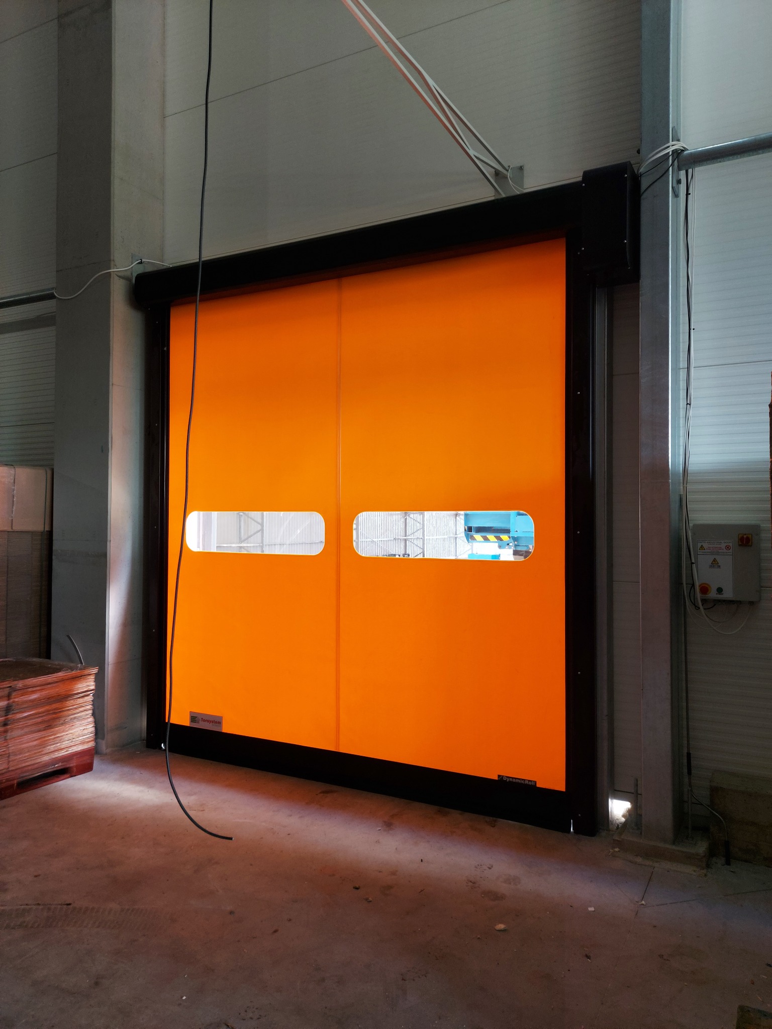 Pomarańczowa, segmentowa kurtyna przemysłowa z dwoma małymi okienkami, widoczna w hali produkcyjnej, z elementami konstrukcyjnymi i przewodami elektrycznymi w tle.