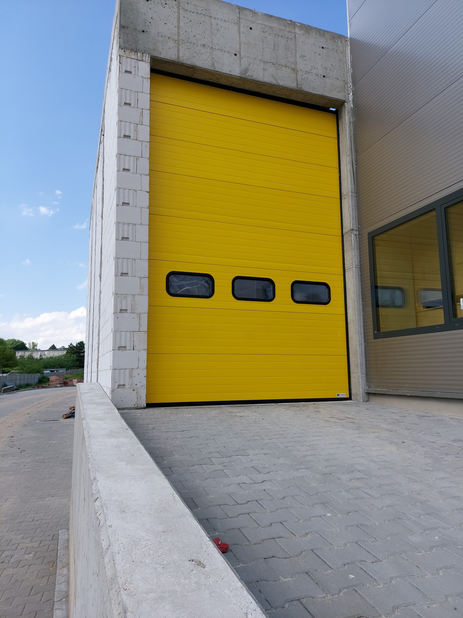 Jasnożółta segmentowa brama z trzema poziomymi okienkami, zamontowana w szarej betonowej konstrukcji budynku przemysłowego, widok z zewnątrz na podjazd z kostki brukowej.