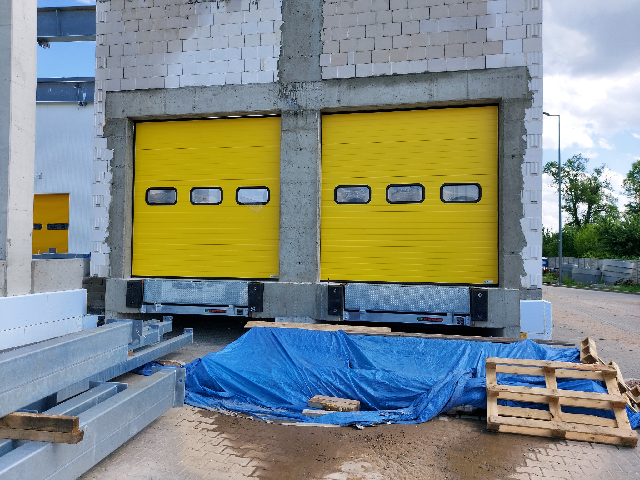 Dwie żółte bramy przemysłowe z czterema prostokątnymi oknami w surowym stanie budynku, z widocznymi elementami konstrukcyjnymi i materiałami budowlanymi na zewnątrz.