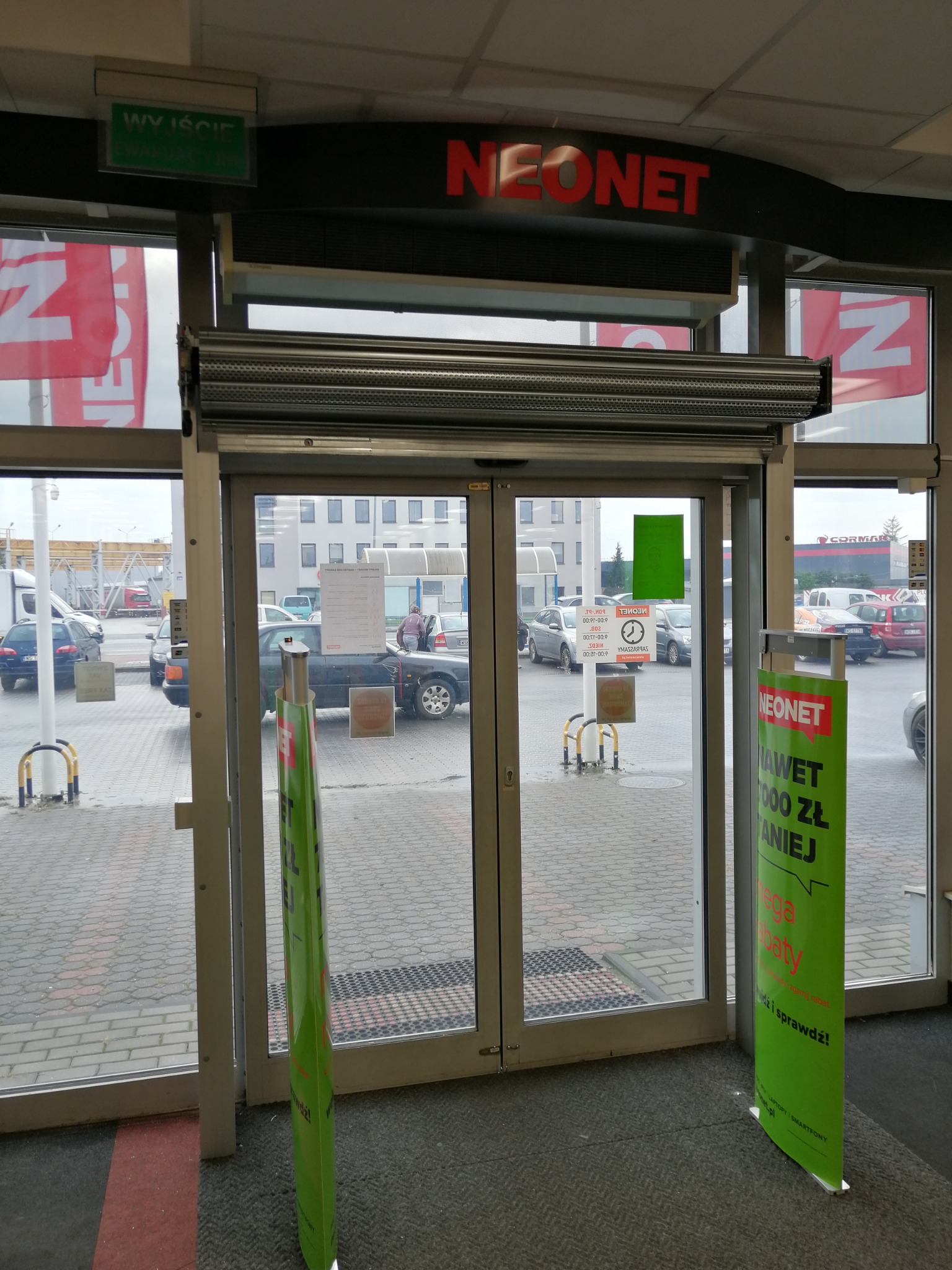 Wejście do sklepu z częściowo opuszczoną metalową roletą zewnętrzną nad drzwiami, widoczne reklamy i parking na zewnątrz.