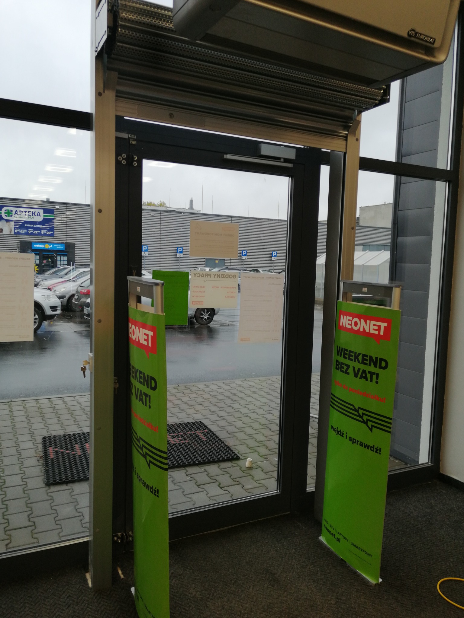 Zewnętrzna roleta antywłamaniowa częściowo opuszczona nad wejściem do sklepu, widok z wnętrza, na zewnątrz widoczne zaparkowane samochody i szyld Apteki.