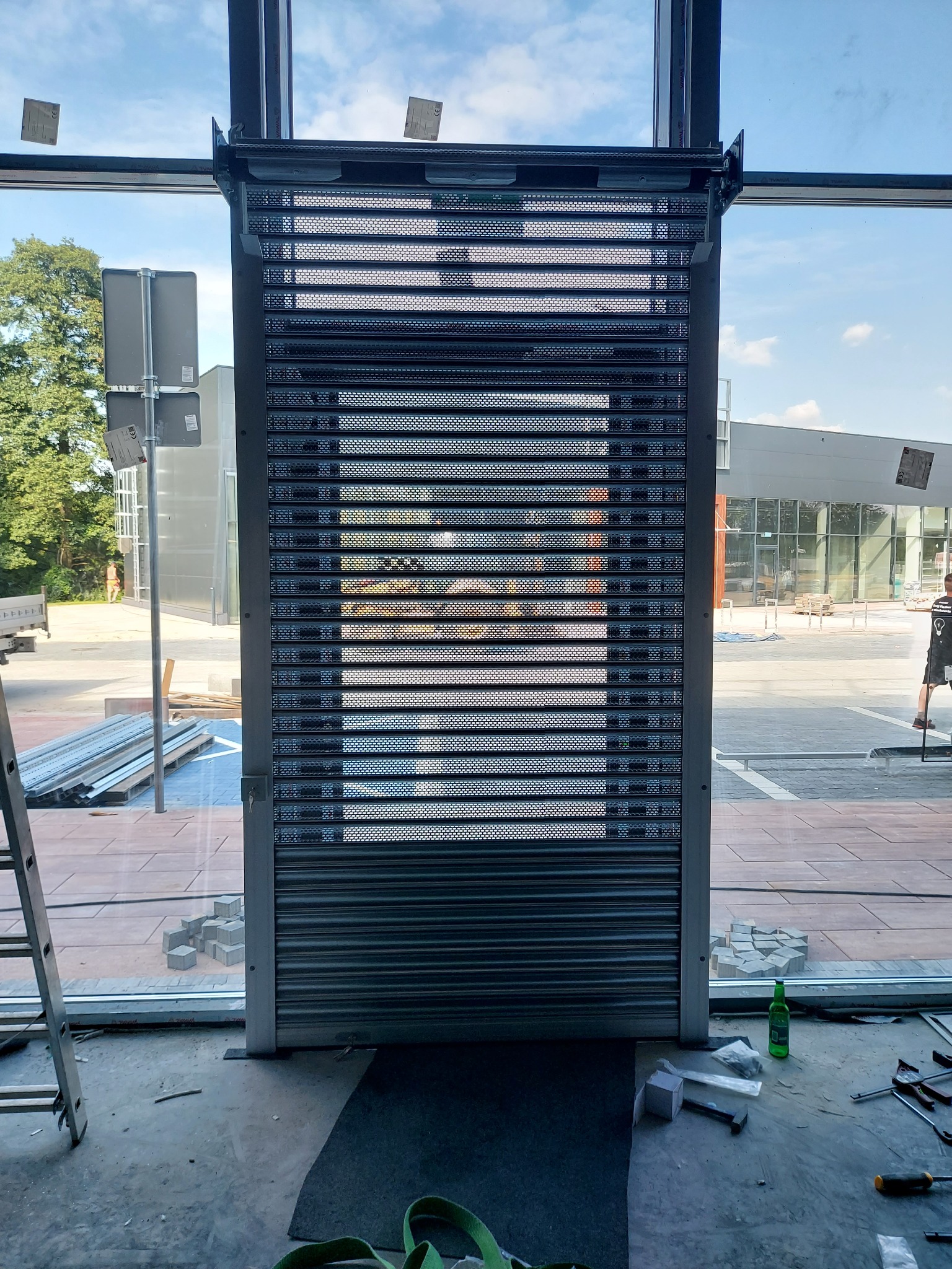 Montaż perforowanej metalowej osłony okiennej w nowym budynku, widoczne narzędzia, butelka i kostka brukowa wewnątrz, plac budowy na zewnątrz.