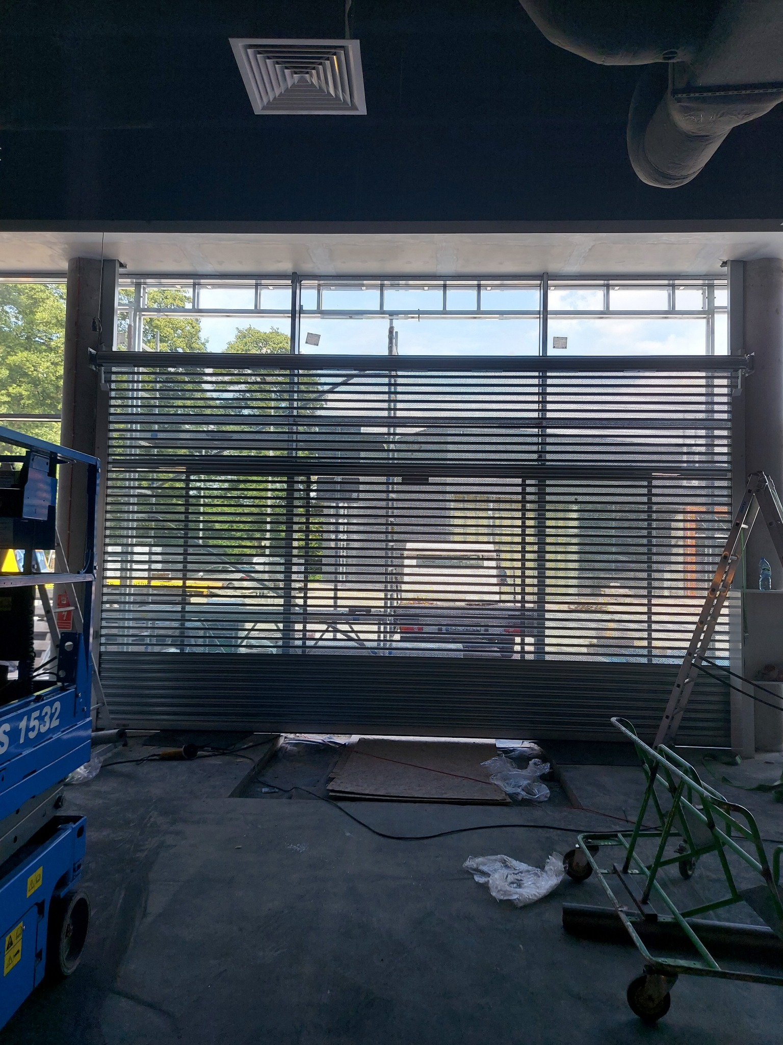 Montaż perforowanej, metalowej rolety w oknie budynku przemysłowego, widoczny podnośnik koszowy i drabina, surowy stan wykończenia wnętrza.