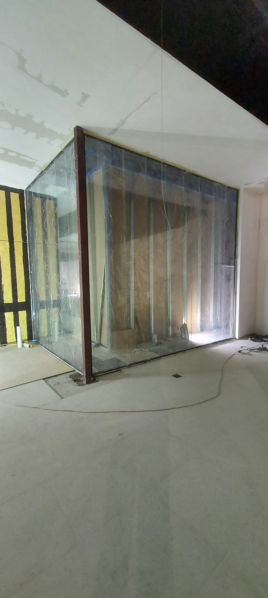 Wnętrze w trakcie prac wykończeniowych, widoczna konstrukcja z profili metalowych osłonięta folią, fragment ocieplonej ściany z wełny mineralnej, surowa podłoga betonowa.