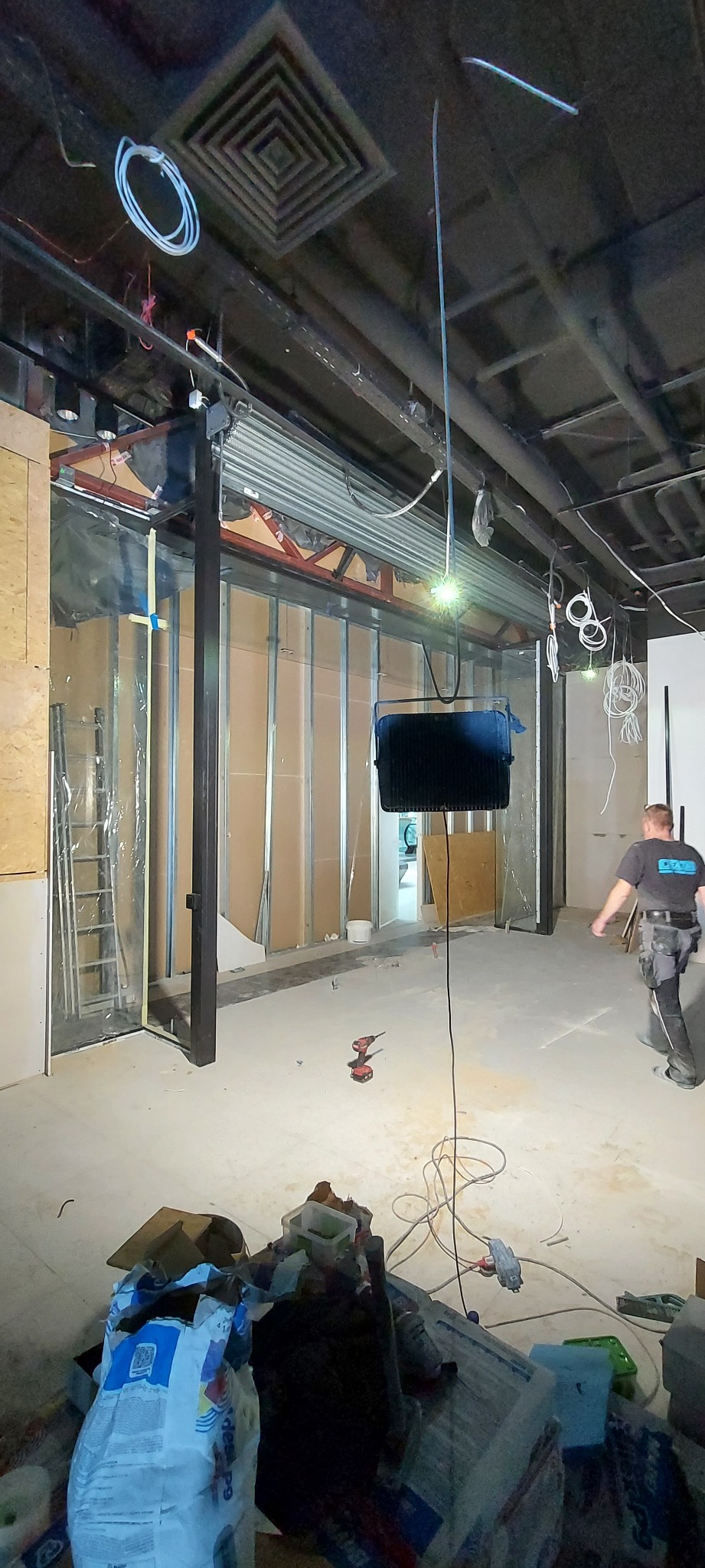 Montaż stalowej konstrukcji pod system roletowy w surowym wnętrzu; widoczne narzędzia, przewody elektryczne, drabina i fragment sylwetki pracownika.