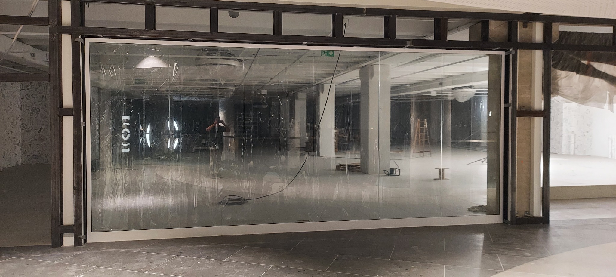 Przeszklona brama przemysłowa z widocznym wnętrzem budynku w trakcie remontu, zabezpieczona folią, z widocznym odbiciem osoby robiącej zdjęcie.