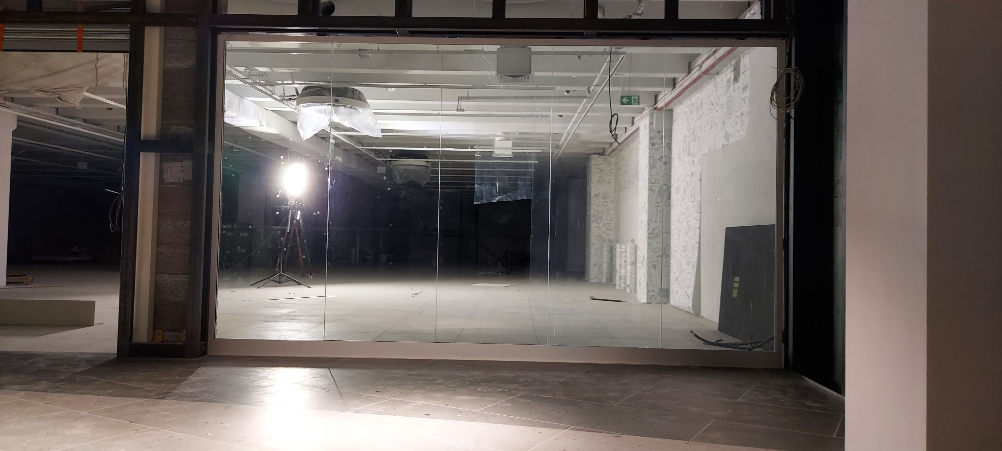 Szklana brama przemysłowa w trakcie montażu, widoczne oświetlenie budowlane i surowy stan wykończenia wnętrza.