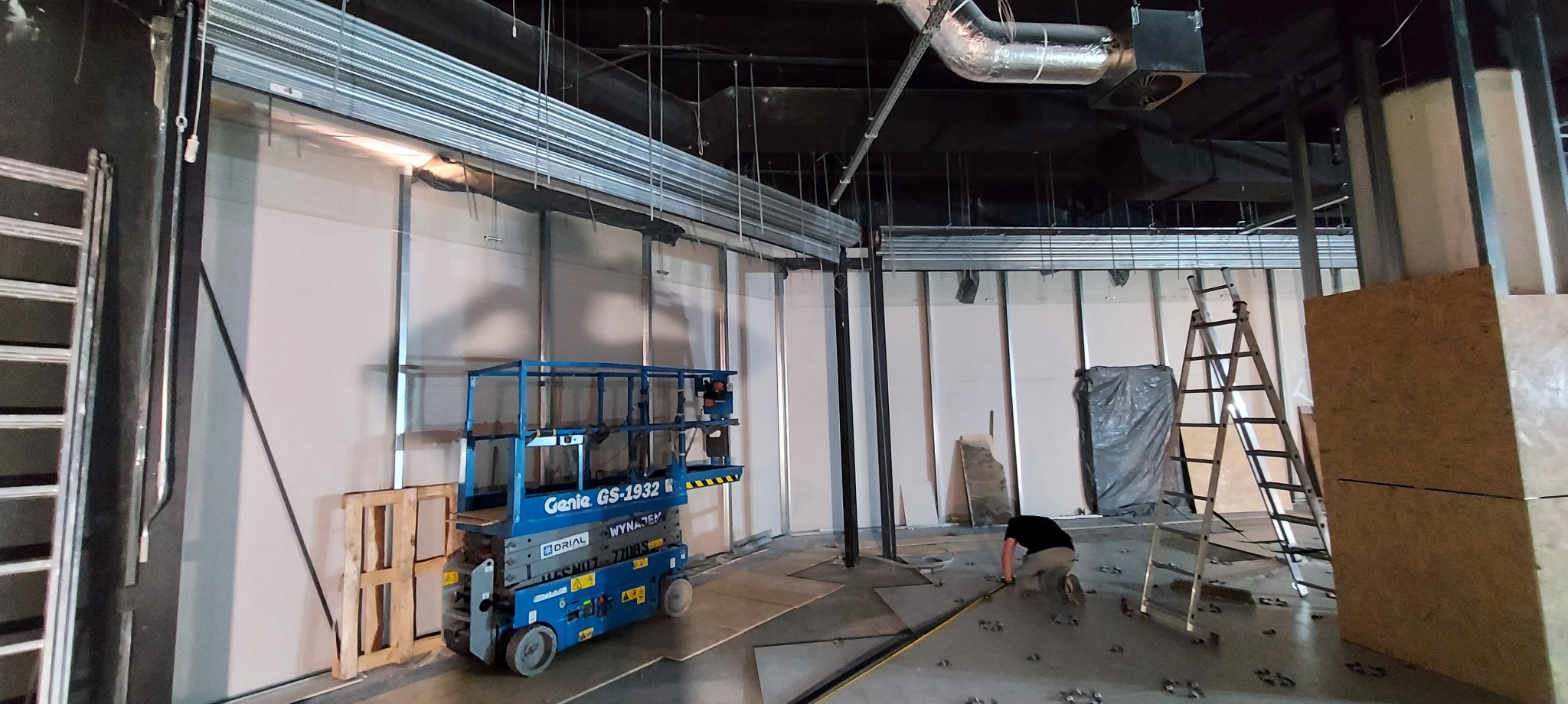 Wnętrze hali w trakcie montażu systemu szynowego pod rolety, widoczny podnośnik nożycowy Genie GS-1932 i pracownik na podłodze mocujący elementy konstrukcyjne.
