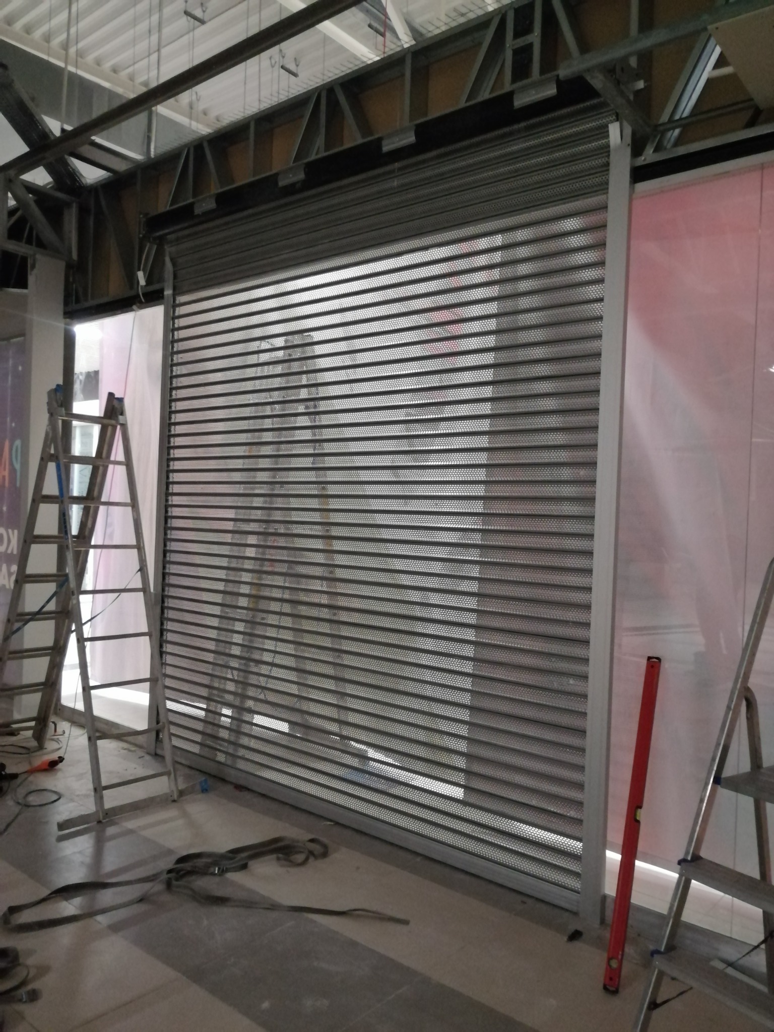 Montaż perforowanej, metalowej rolety w surowym wnętrzu, widoczne drabiny, poziomica i elementy konstrukcyjne budynku.