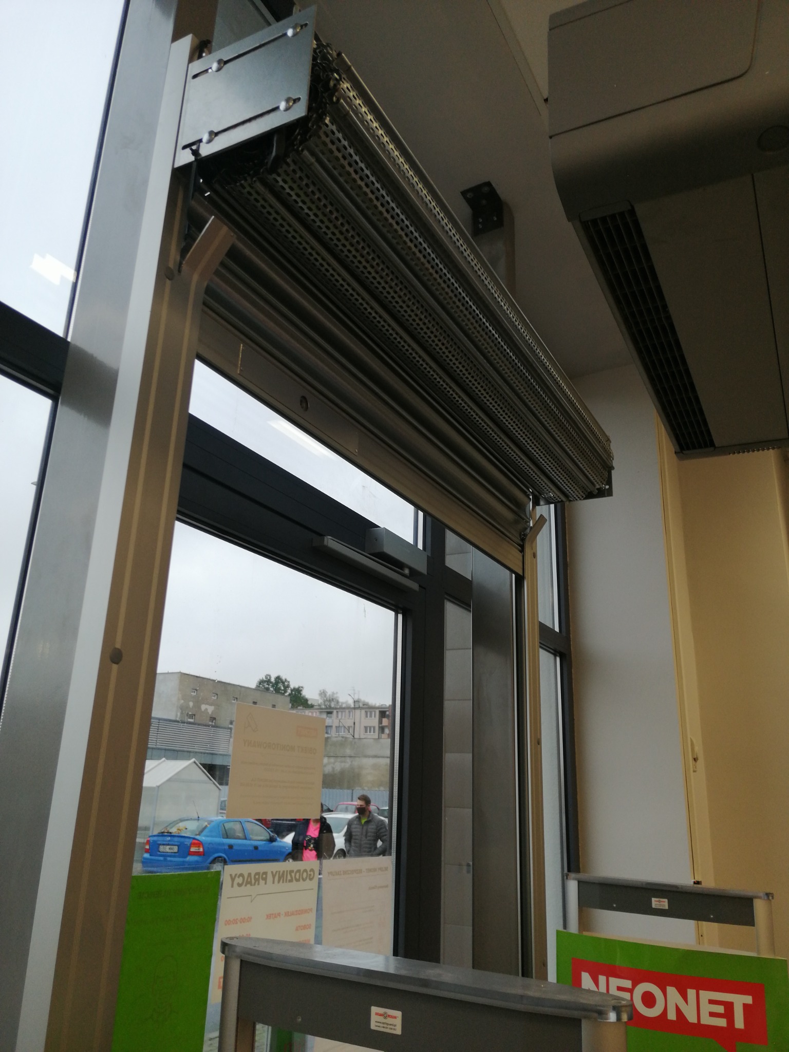 Zamontowana metalowa roleta perforowana, częściowo opuszczona, widoczna z perspektywy wewnętrznej, z widokiem na ulicę i zaparkowany niebieski samochód.