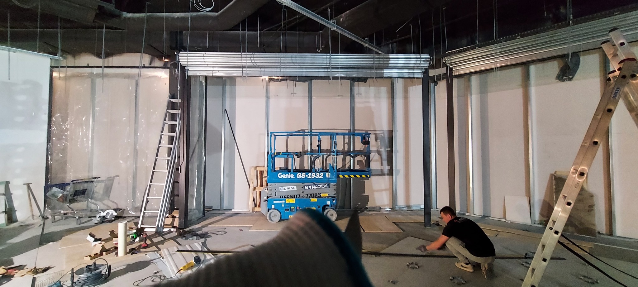 Montaż metalowej konstrukcji pod osłonę, widoczny podnośnik Genie GS-1932 i pracownik podczas układania elementów na podłodze.