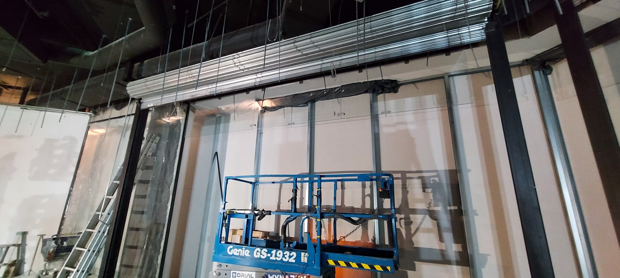 Montaż metalowej rolety przemysłowej w surowym wnętrzu, widoczny podnośnik Genie i konstrukcja ścianek działowych.
