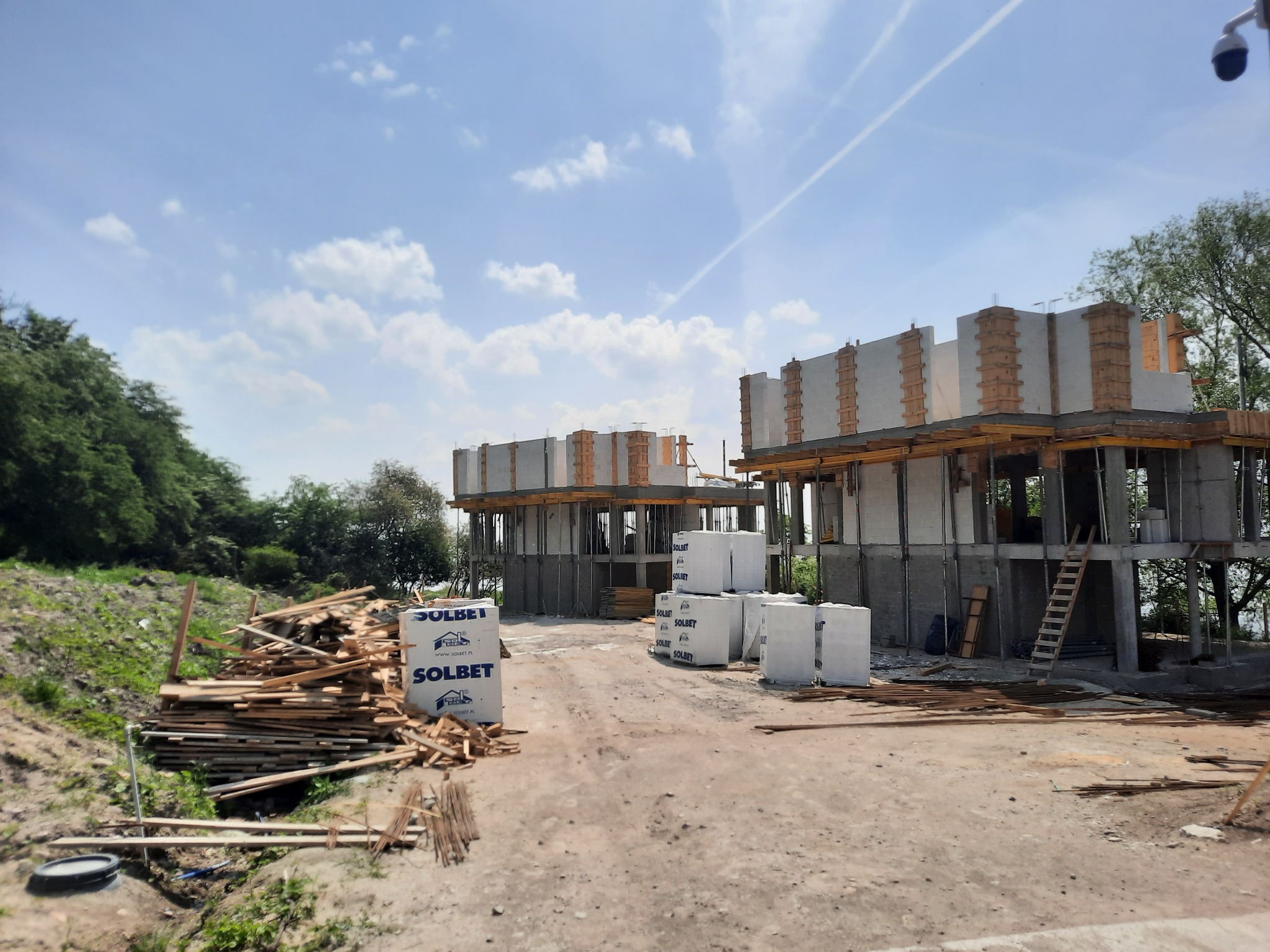 Budowa kompleksu budynków w Lubiniu, widoczne surowe konstrukcje budynków w trakcie wznoszenia, materiały budowlane marki Solbet oraz stos drewna na pierwszym planie.