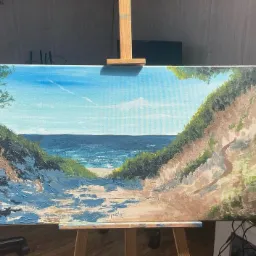 Obraz olejny przedstawiający pejzaż morski z klifami i plażą, umieszczony na drewnianej sztaludze w pomieszczeniu.